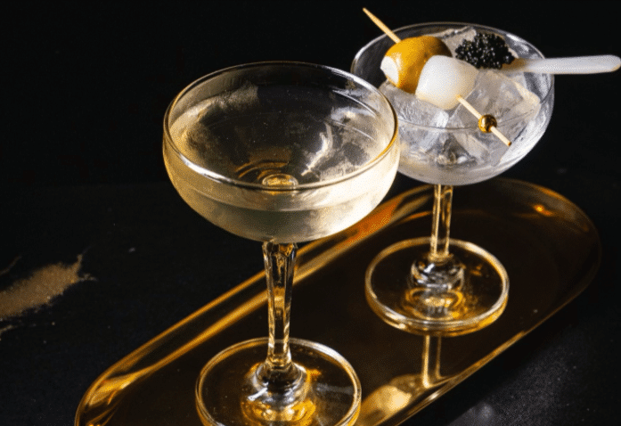 Two elegant coupe glasses on a golden tray; one filled with a clear drink, the other with ice, garnished with olives, a lemon twist, and caviar on a pick, against a dark background.