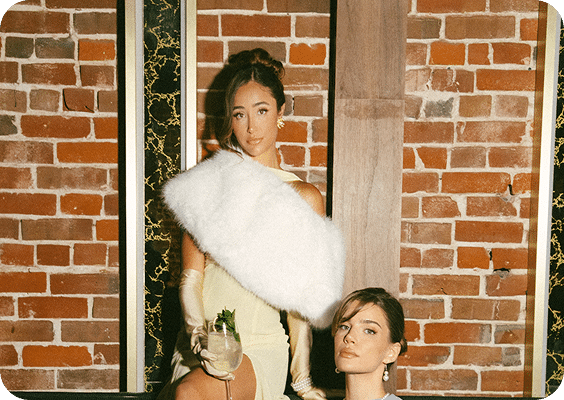 Two women dressed formally pose in front of a brick wall. One woman stands wearing a light dress, long gloves, and a white fur stole, holding a cocktail. The other woman sits beside her, both looking toward the camera.