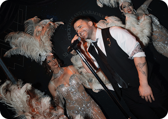 A man in a vest and tie sings into a microphone on stage, smiling. He is surrounded by performers in elaborate feathered costumes and headpieces, creating a festive, cabaret-style atmosphere.