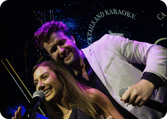 A smiling man and woman stand close together, each holding a microphone while singing karaoke. The background features a dark, starry design with text that includes "Cocktails and Karaoke.