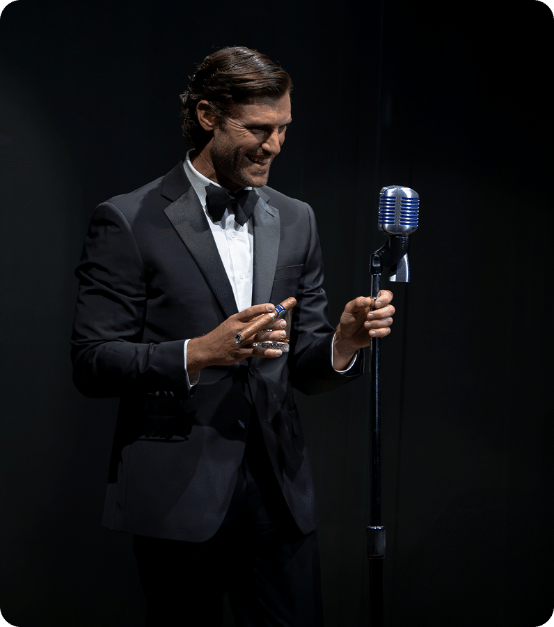 A man in a black tuxedo smiles while holding a drink and a microphone on a stand, standing against a dark background.