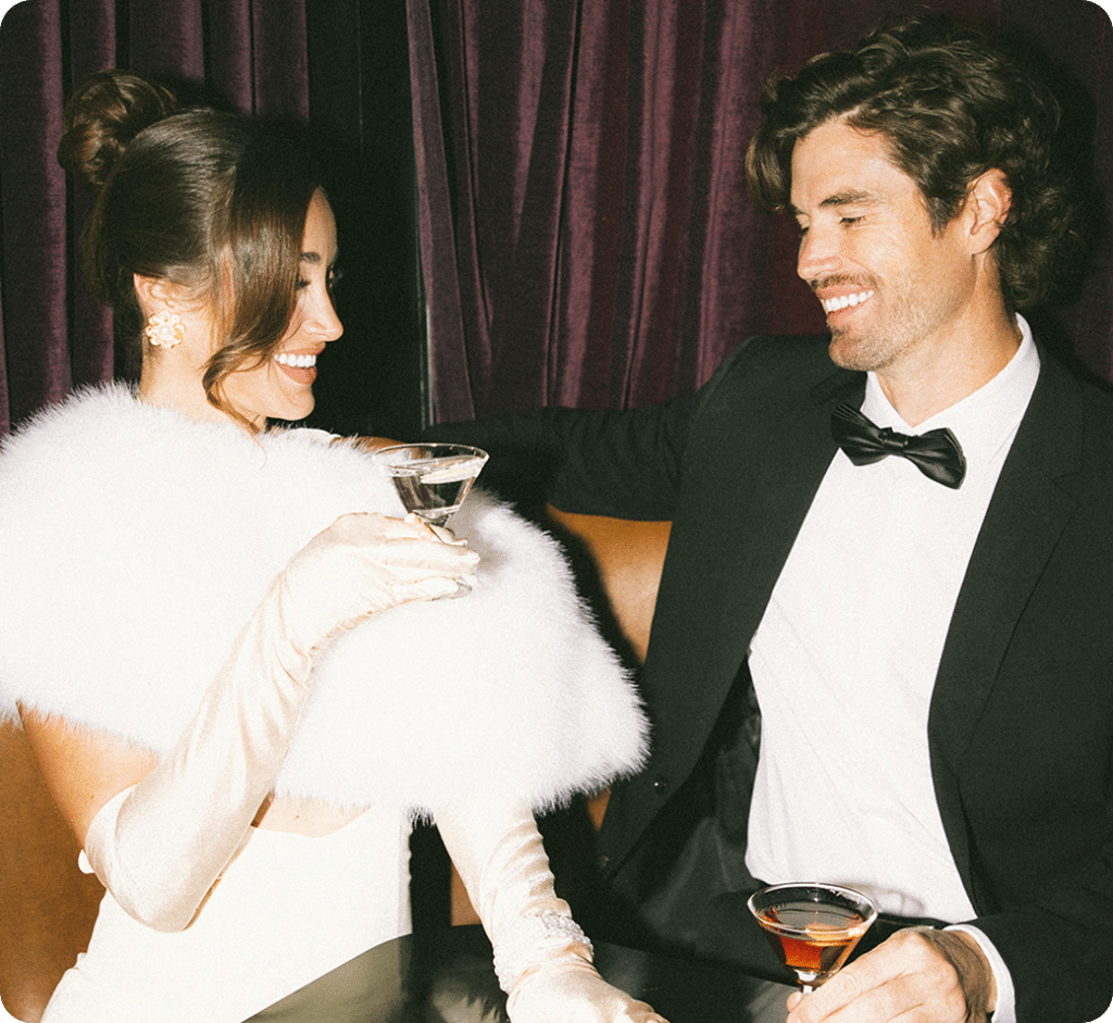 A woman in an elegant white dress and gloves holds a cocktail and smiles at a man in a black tuxedo. They sit together on a brown sofa in front of deep purple curtains, appearing happy and engaged in conversation.