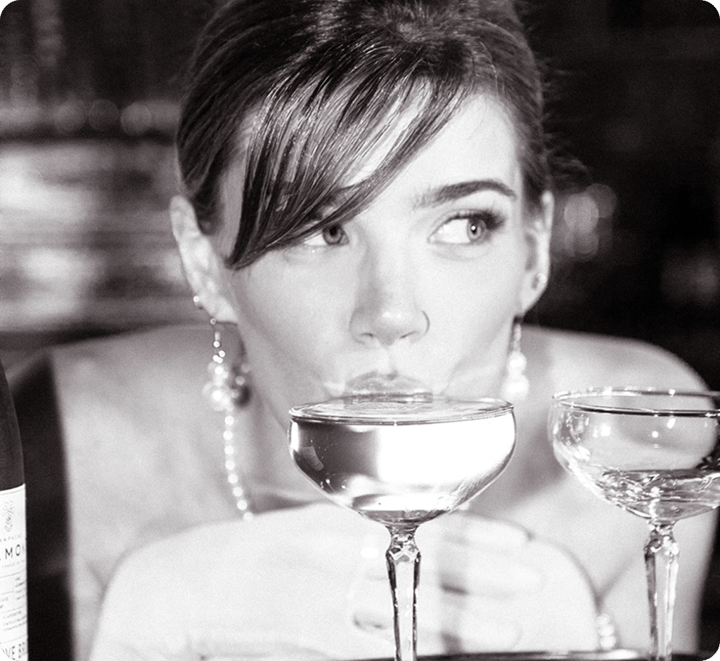 A woman with earrings and an updo hairstyle sips from a coupe glass, with another empty coupe glass beside her. The image is in black and white and has a glamorous, vintage feel.