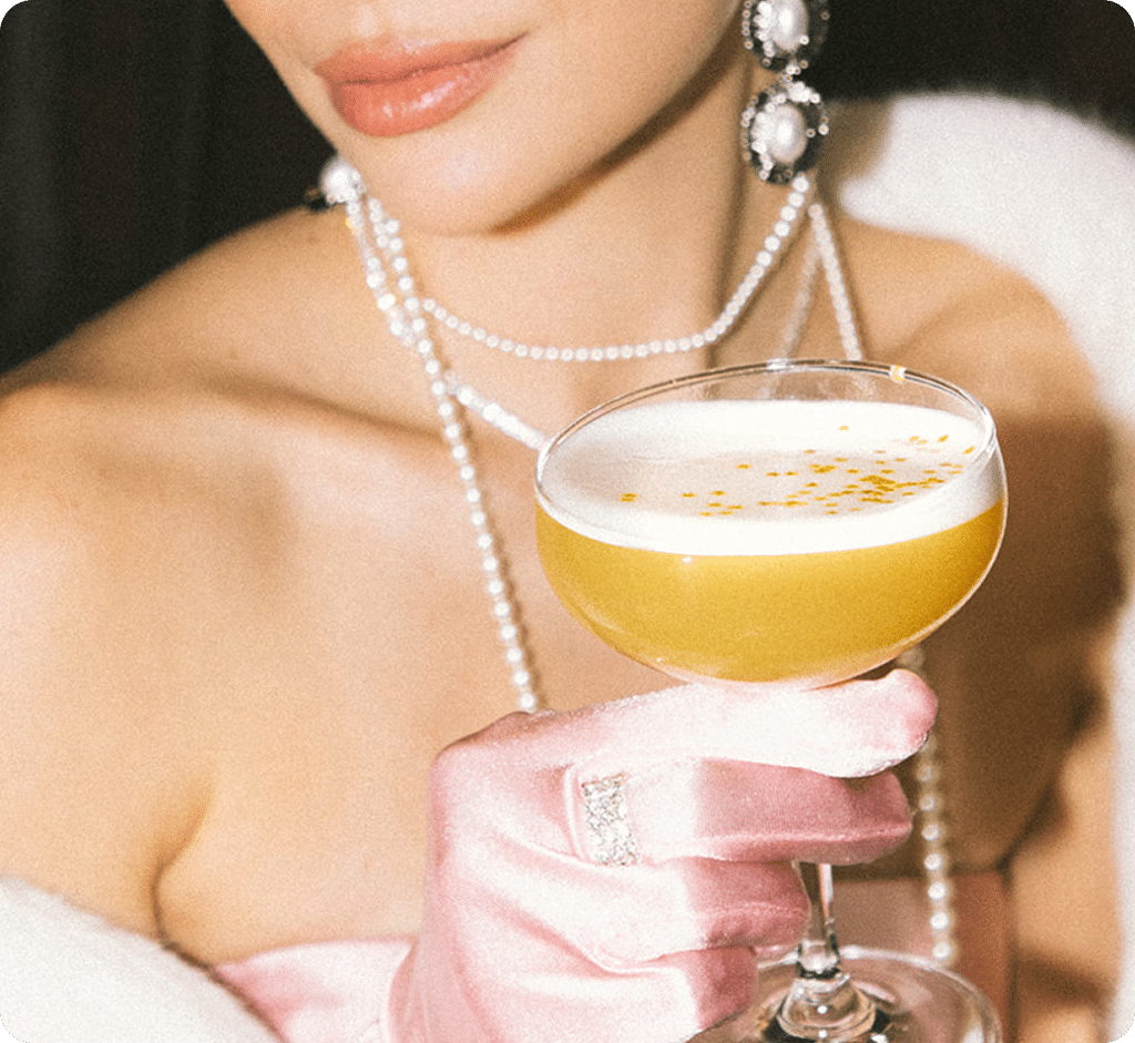 A woman wearing pearls, statement earrings, and a white fur stole holds a coupe glass with a yellow cocktail topped with foam and gold sprinkles. She wears pink satin gloves and a diamond ring.