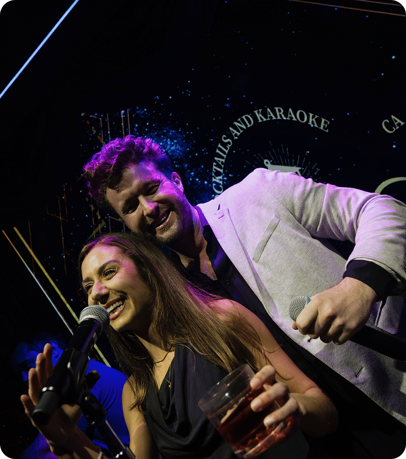Two people are smiling and singing into microphones on stage. The woman in front holds a drink, and the man behind her wears a light-colored blazer. The background has neon lights and a sign mentioning "cocktails and karaoke.