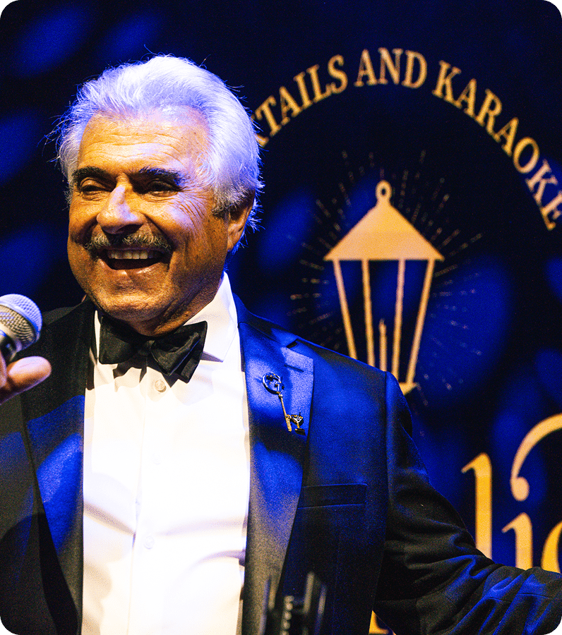 A smiling man with gray hair in a tuxedo stands at a microphone on stage, with a sign behind him that reads "Cocktails and Karaoke" and a lantern graphic.