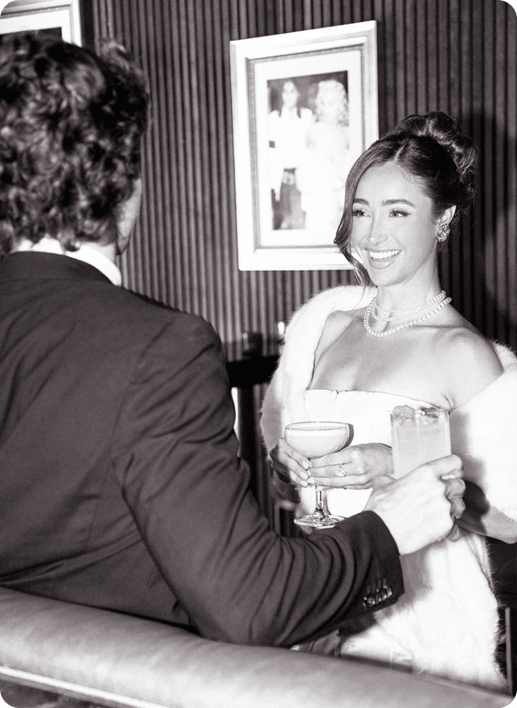A woman in an elegant dress and pearl necklace smiles while holding two cocktails, speaking with a man in a suit at a stylish bar. Framed photos decorate the wall behind them. The image is in black and white.
